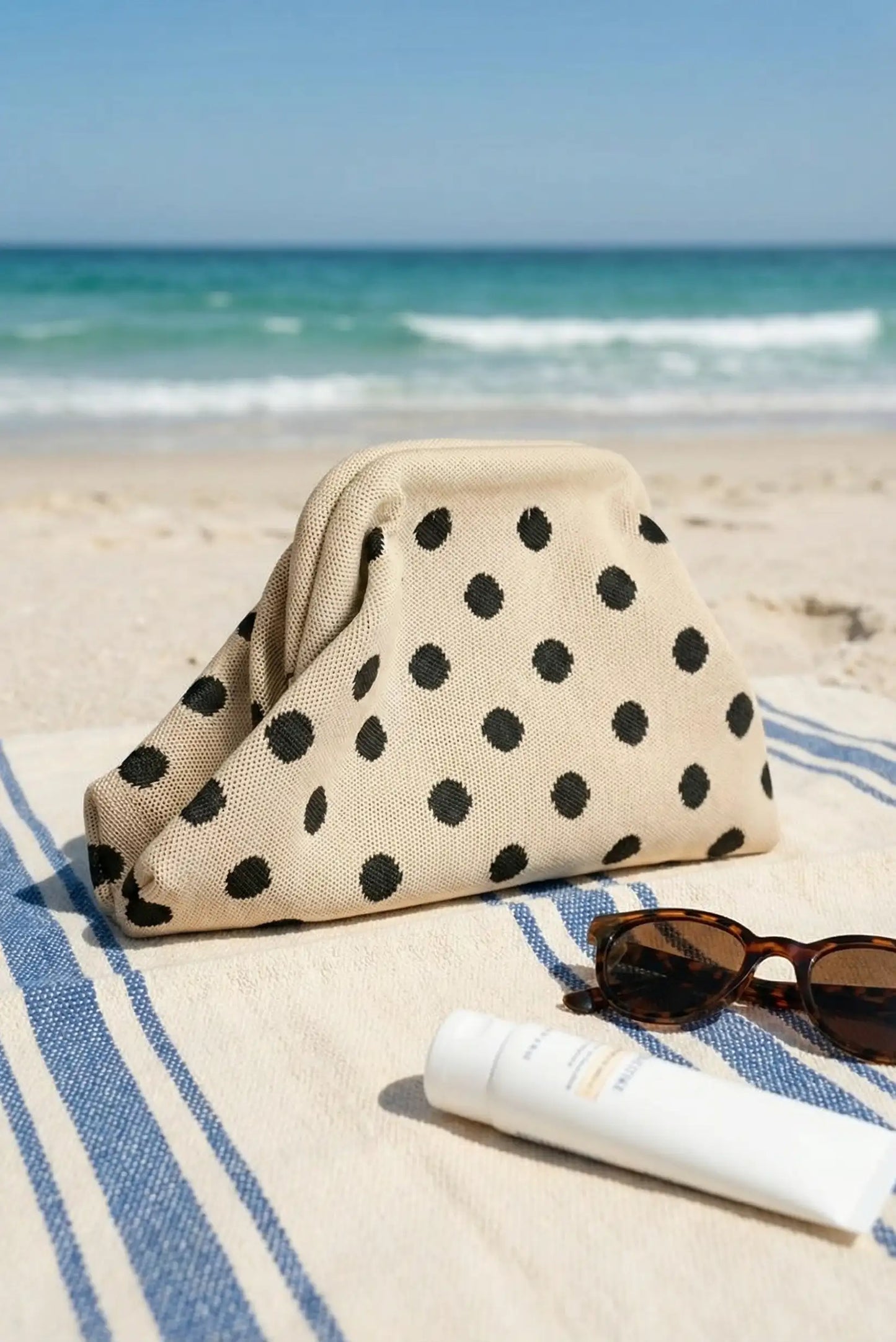 Bolso clutch beige de topos negros sobre toalla en la playa, junto a gafas y crema solar