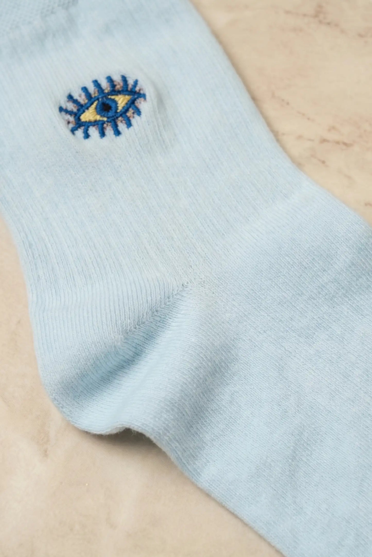Calcetín azul claro de mujer con bordado de ojo turco, sobre fondo beige.
