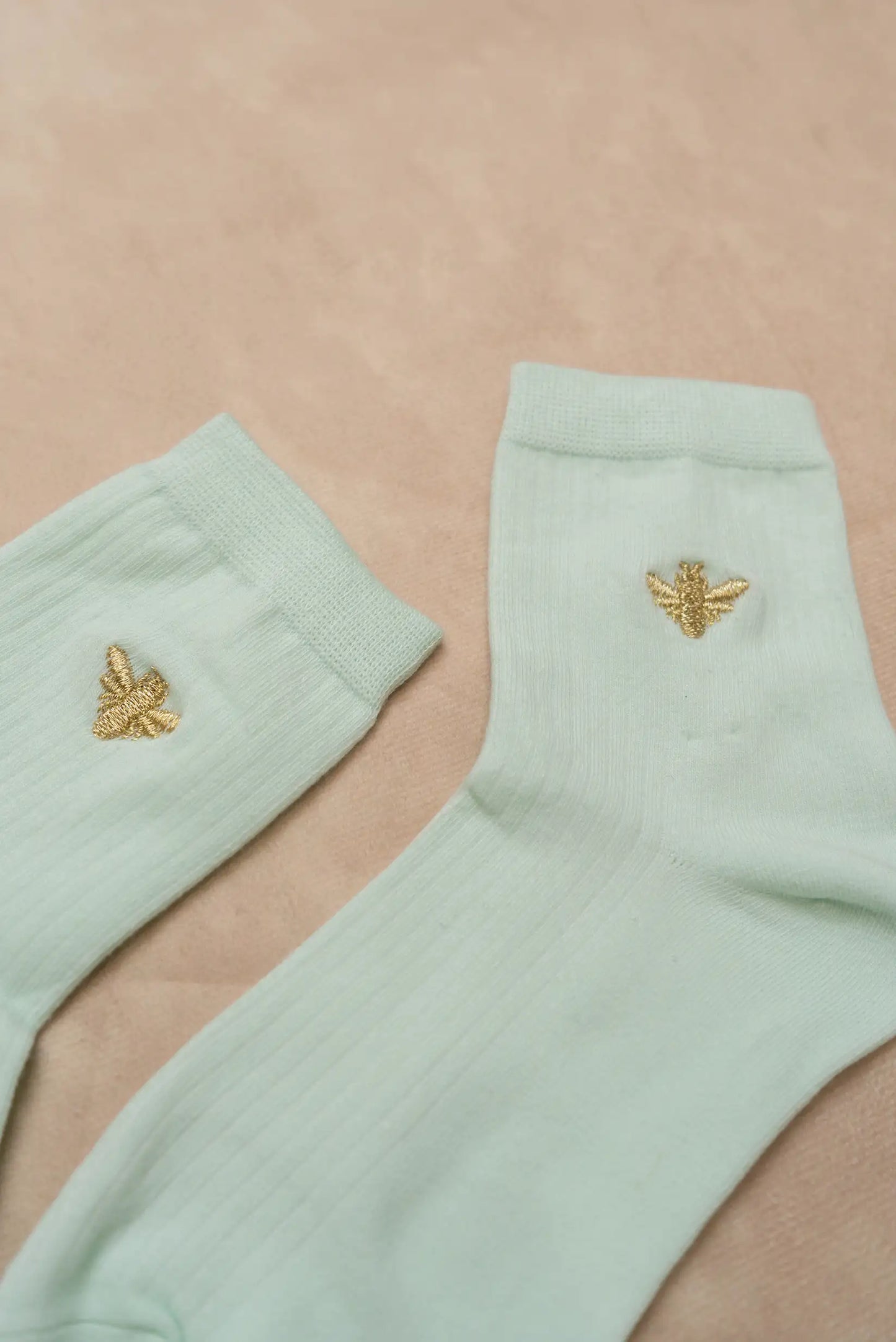 Calcetines de mujer color verde claro con detalle bordado de abeja dorada, fondo beige.