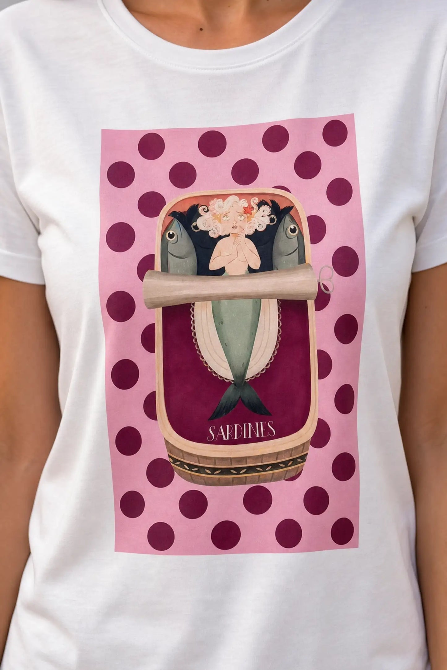 Camiseta blanca con diseño de sirena y sardinas en lata, fondo rosa con lunares