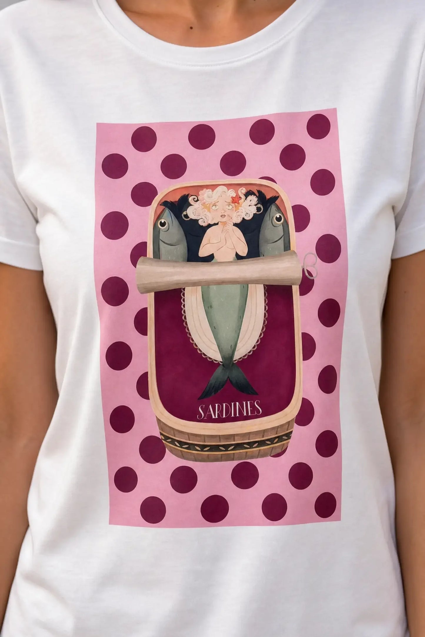 Camiseta blanca con diseño de sirena y sardinas en lata, fondo rosa con lunares