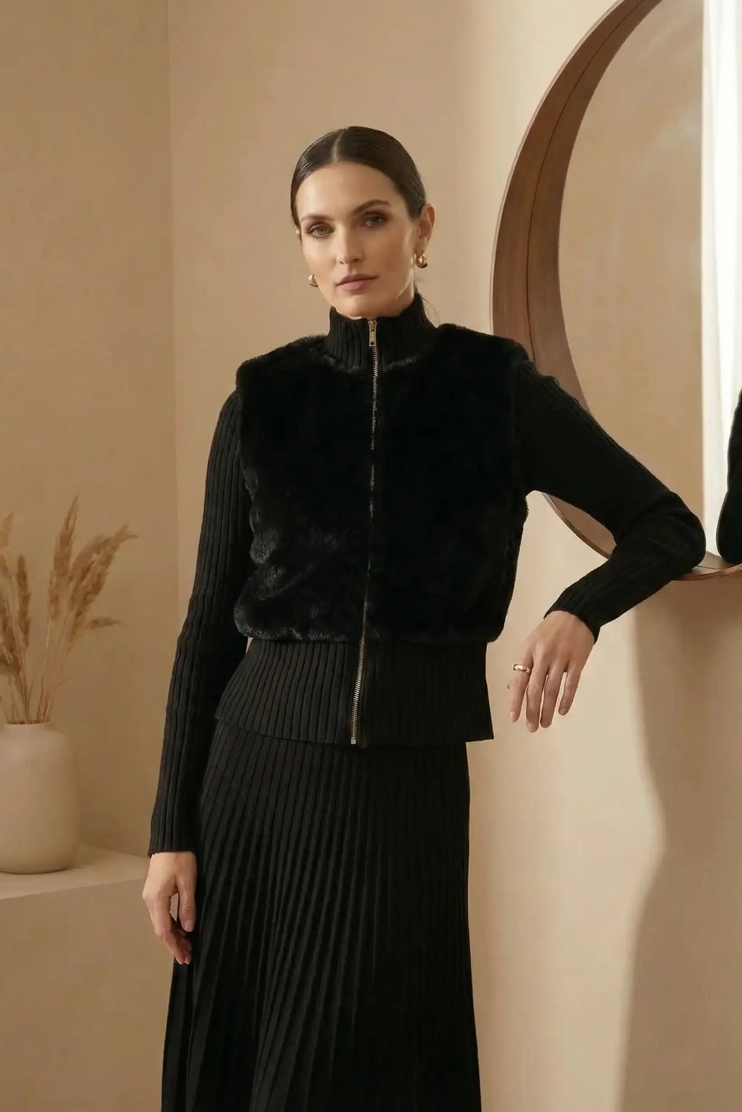 Mujer con chaqueta negra de pelo sintético y falda negra de punto frente a espejo redondo