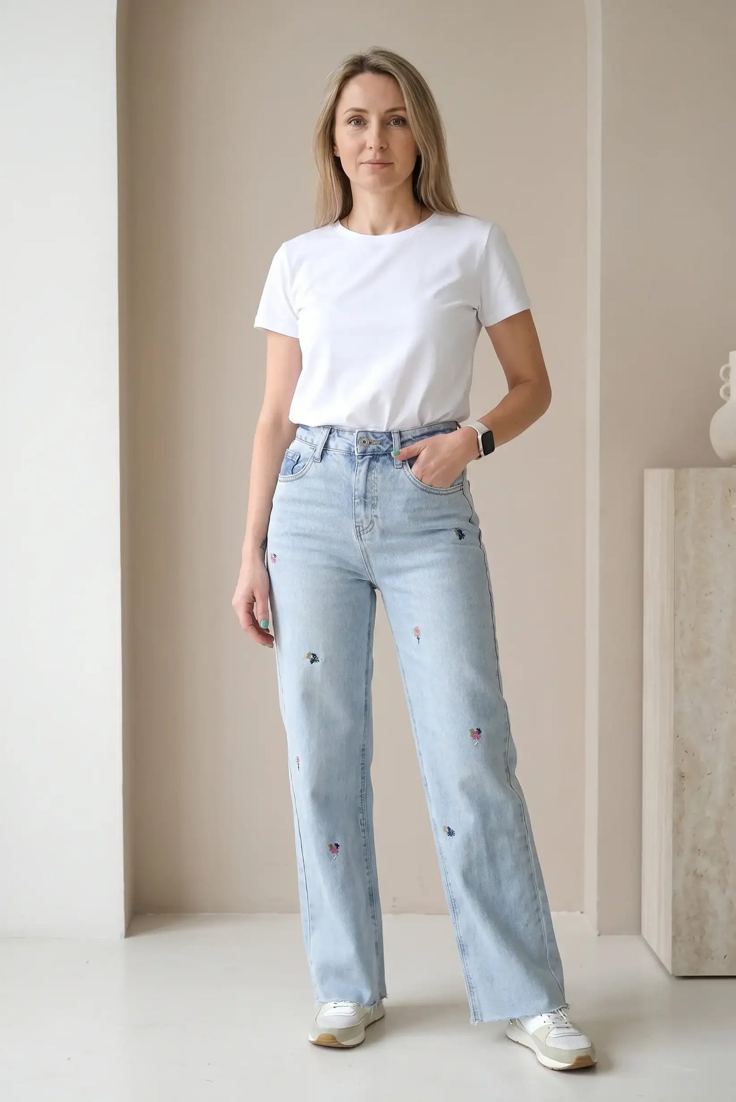 Mujer con camiseta blanca y pantalón vaquero largo azul claro con bordados de flores.