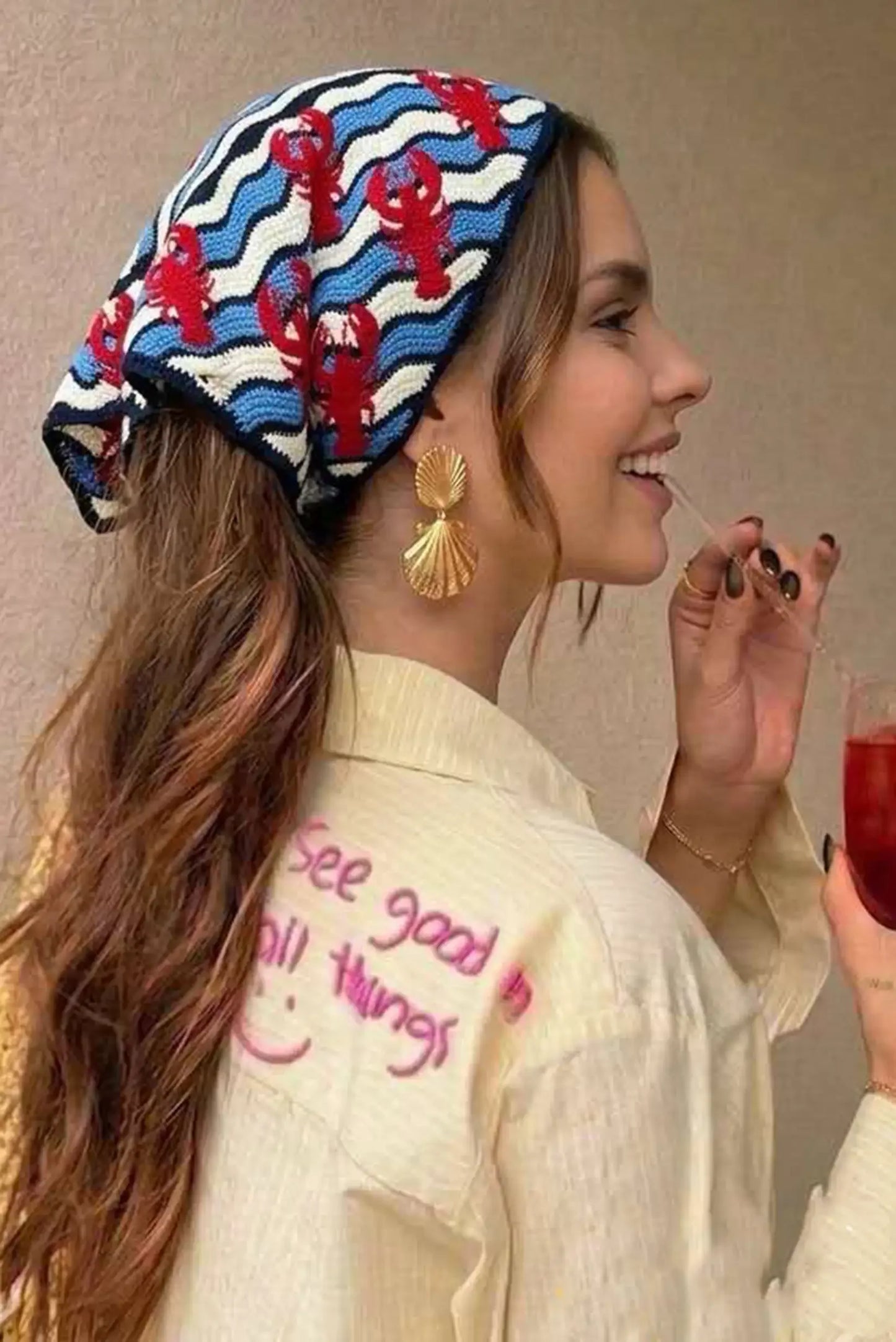 Mujer con gorro tejido de langosta y olas, pendientes de concha y camisa con mensaje 'See good in all things'