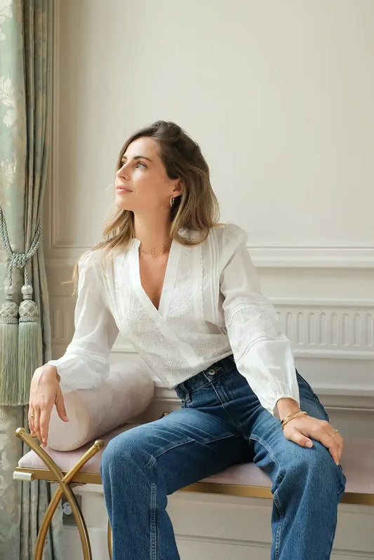 Mujer con camisa blanca bordada de manga larga y jeans sentada en sillón elegante