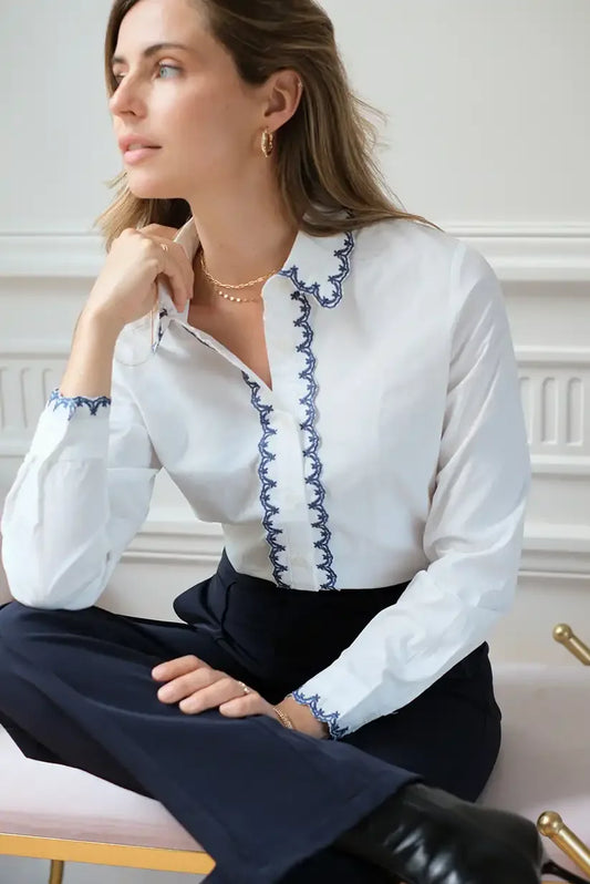 Mujer con camisa blanca de manga larga con detalles en azul, sentada elegantemente en interior.