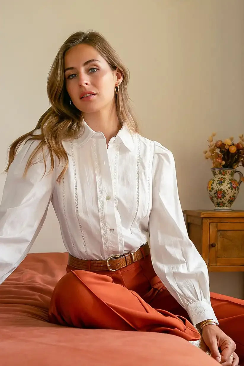 Mujer con camisa blanca de manga larga sentada, pantalón naranja, fondo interior acogedor