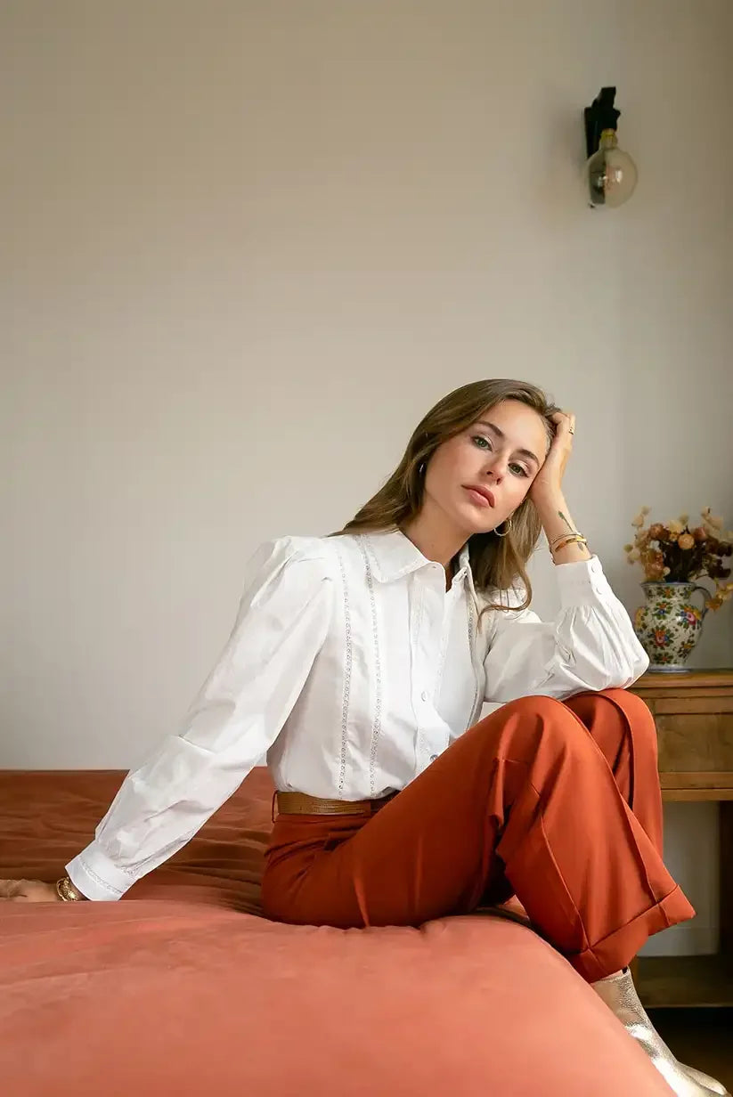 Mujer con camisa blanca manga larga cuello mao y pantalón naranja sentada en cama