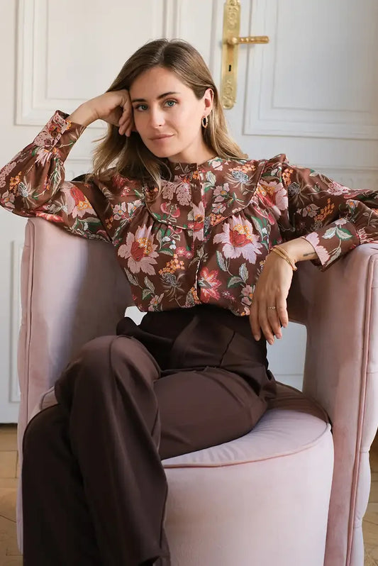 Mujer sentada con camisa marrón de flores otoñales y pantalón marrón en sillón rosa.