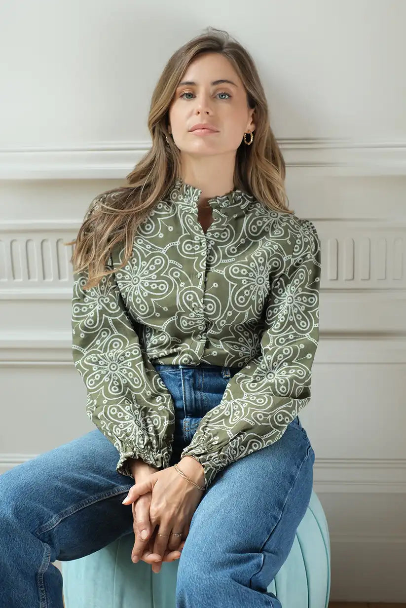 Mujer con camisa verde de manga larga y bordados florales blancos, sentada en interior elegante.