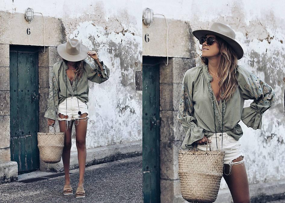 Mujer con look boho, blusa verde, shorts blancos, sombrero y bolso de mimbre frente a pared rústica