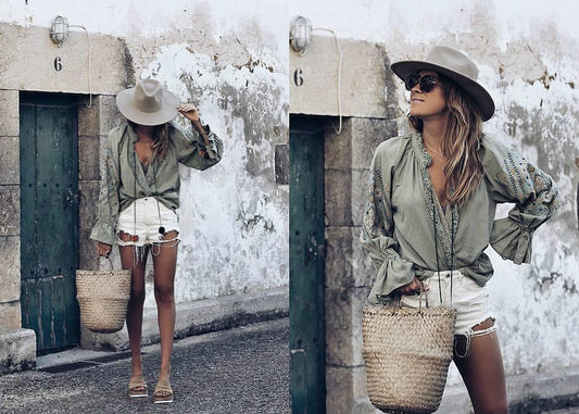 Mujer con look boho, blusa verde, shorts blancos, sombrero y bolso de mimbre frente a pared rústica