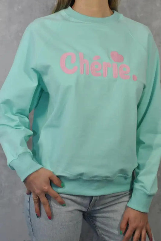 Mujer con sudadera verde agua con texto Chérie rosa, jeans claros, fondo gris.