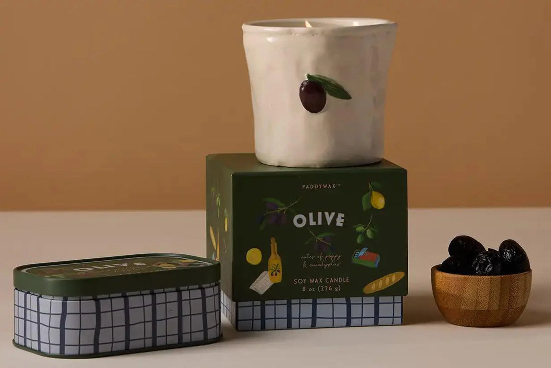 Vela aromática Paddywax Olive en envase cerámico y caja verde, junto a aceitunas negras.