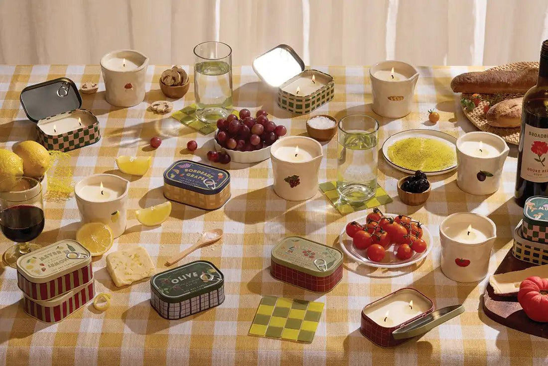 Velas aromáticas Paddywax colección Bistro sobre mesa con mantel a cuadros y comida variada