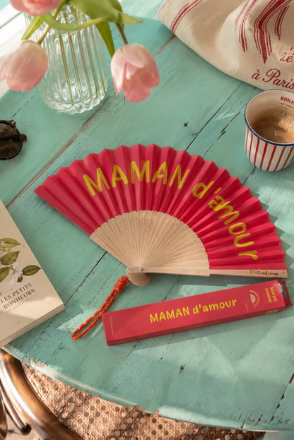 Abanico rosa con texto 'MAMAN d'amour' en mesa turquesa, ideal como regalo para mamá