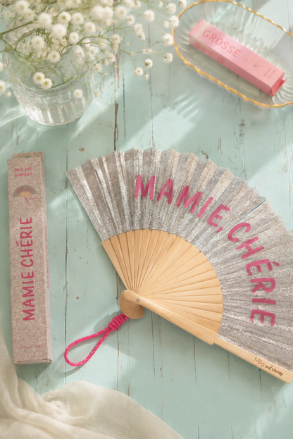 Abanico con texto 'MAMIE CHÉRIE' en rosa, sobre fondo de madera clara con flores y caja de regalo