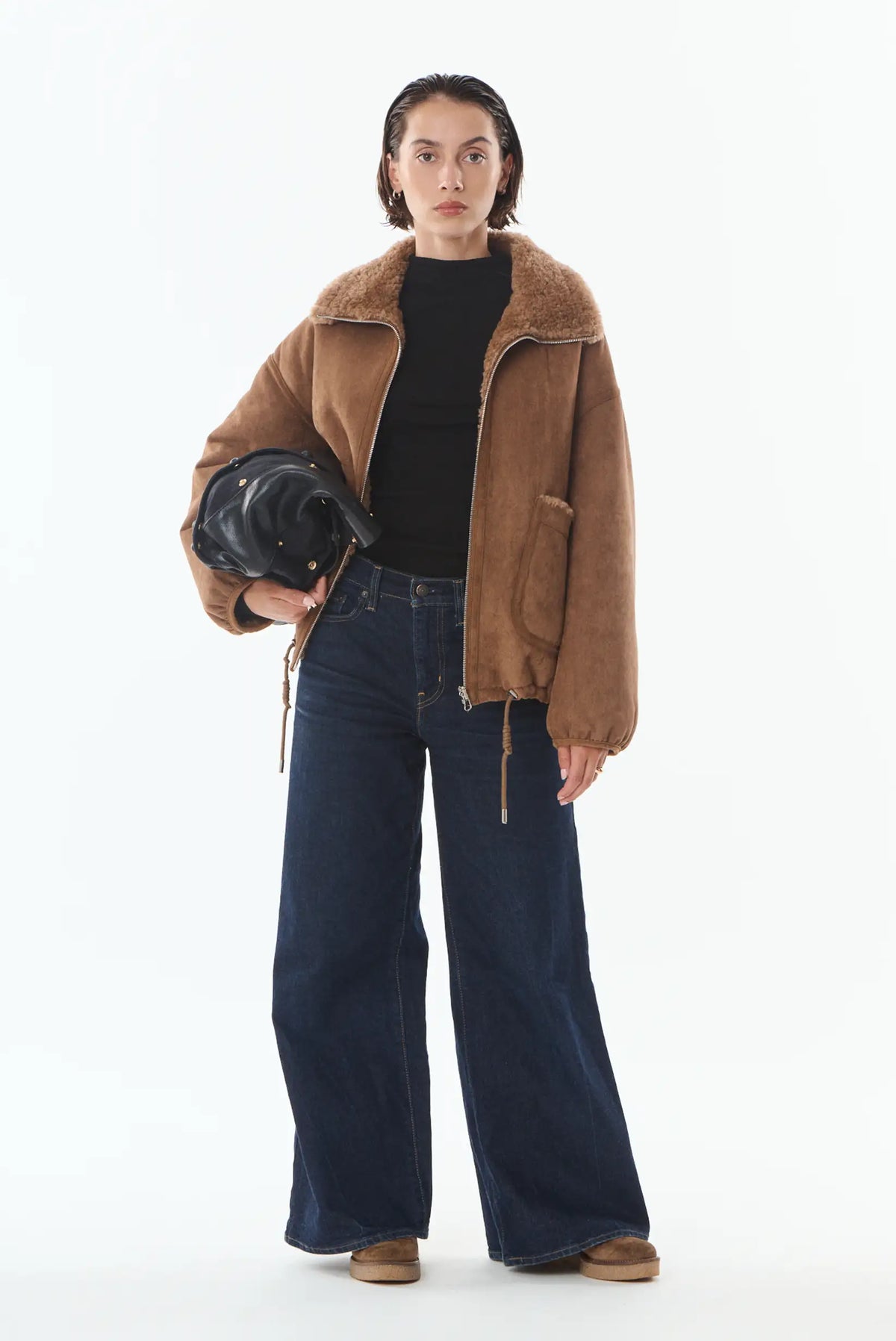 Mujer con chaqueta marrón de borreguillo, vaqueros anchos azules y bolso negro.