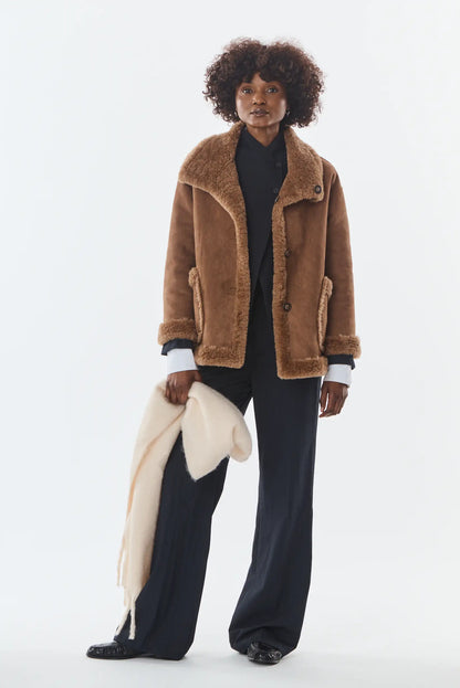 Mujer con chaqueta de ante marrón con borreguillo, pantalón negro y manta beige.