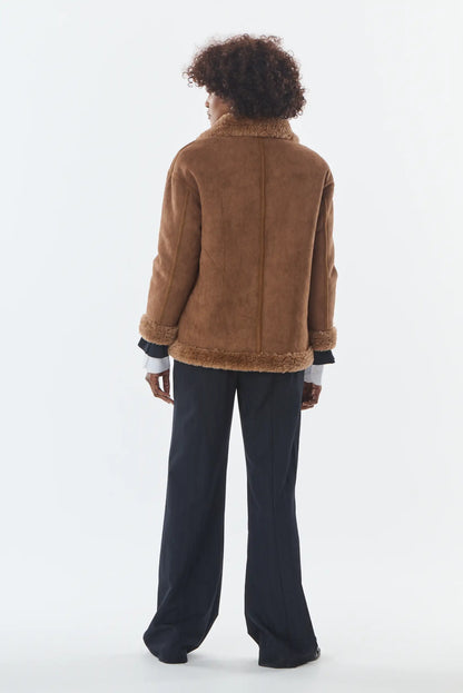 Vista trasera de mujer con chaqueta de ante marrón con forro de borreguito y pantalones negros.