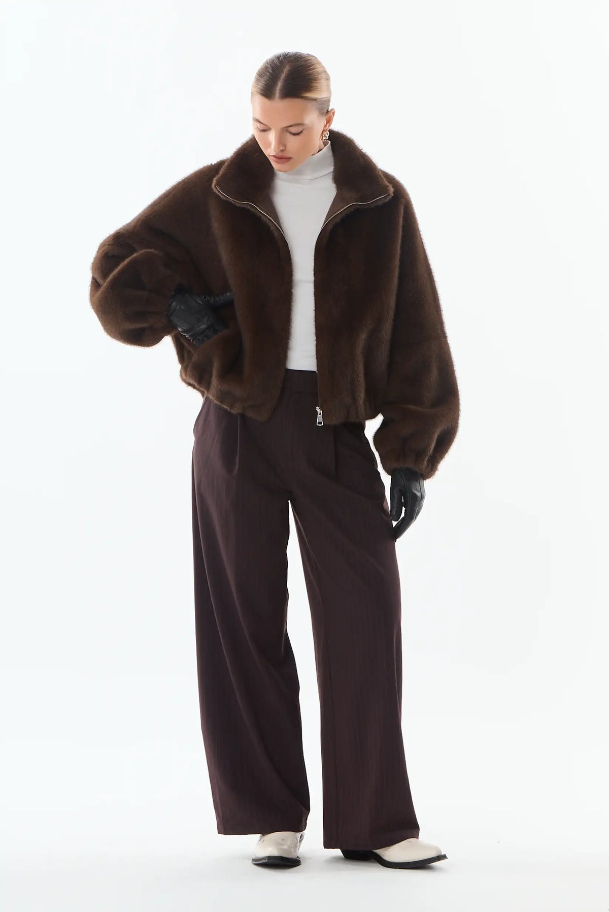 Mujer con chaqueta de piel sintética marrón, jersey blanco y pantalones anchos burdeos.