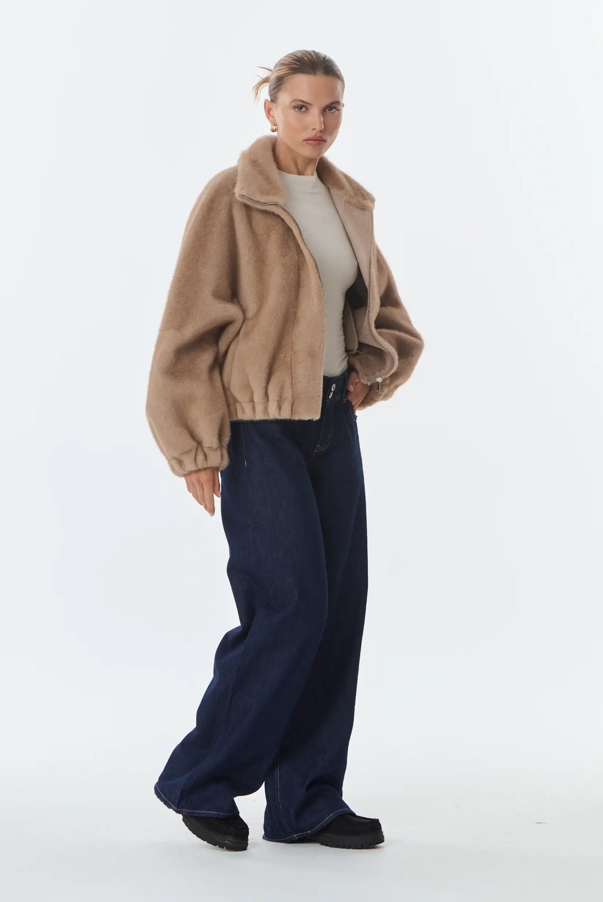 Mujer con chaqueta de efecto piel beige, camiseta blanca y vaqueros anchos azul oscuro.