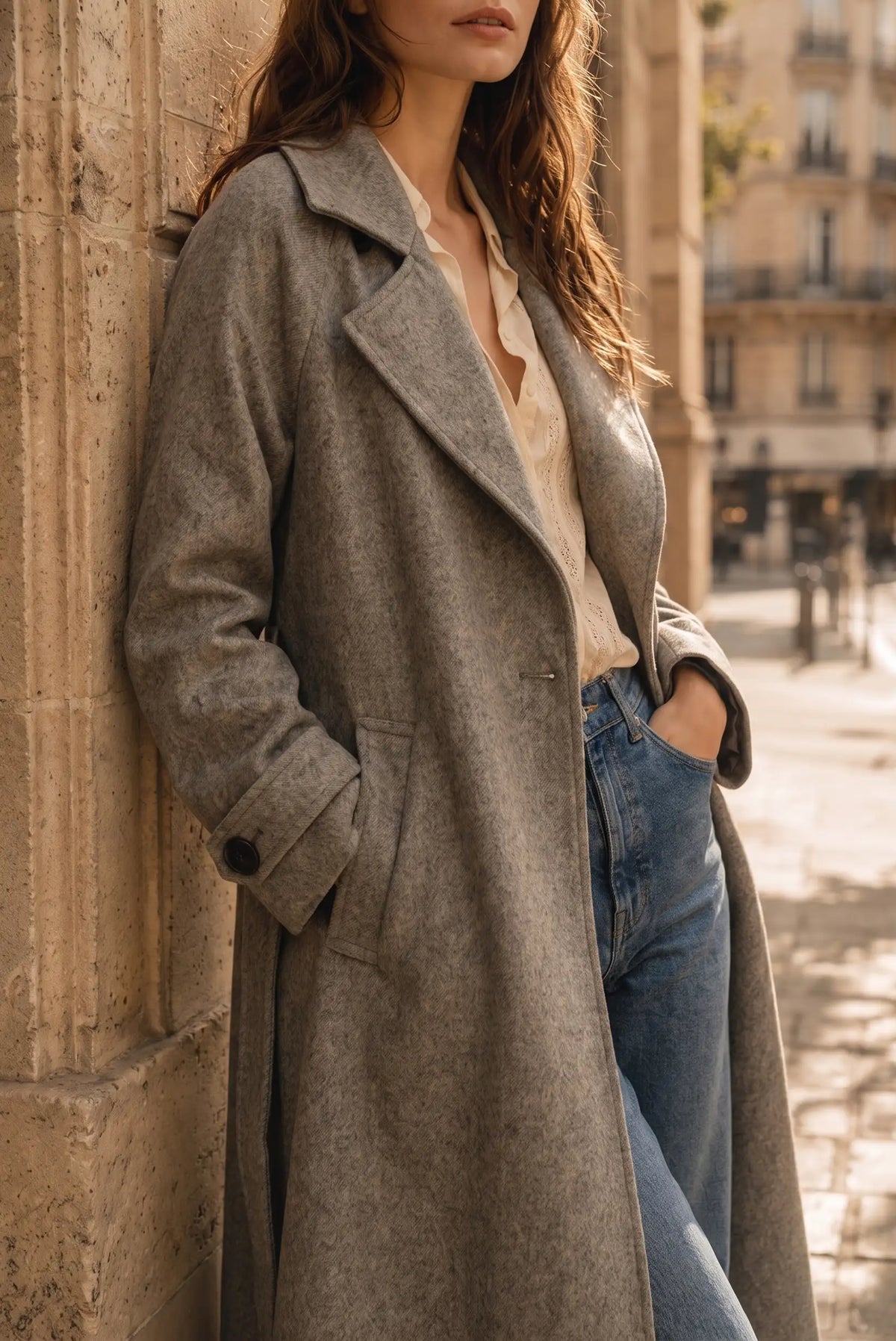 Mujer con abrigo largo gris, blusa beige y vaqueros azules en entorno urbano.