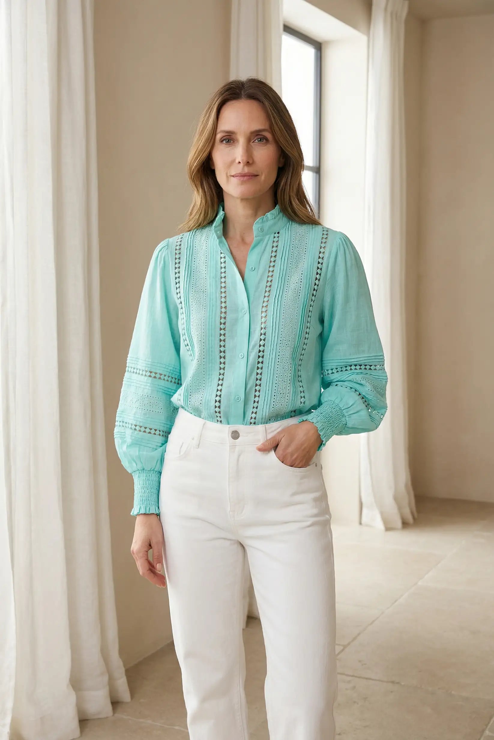 Mujer con blusa verde agua de manga larga y bordados, combinada con pantalón blanco, en ambiente interior luminoso.