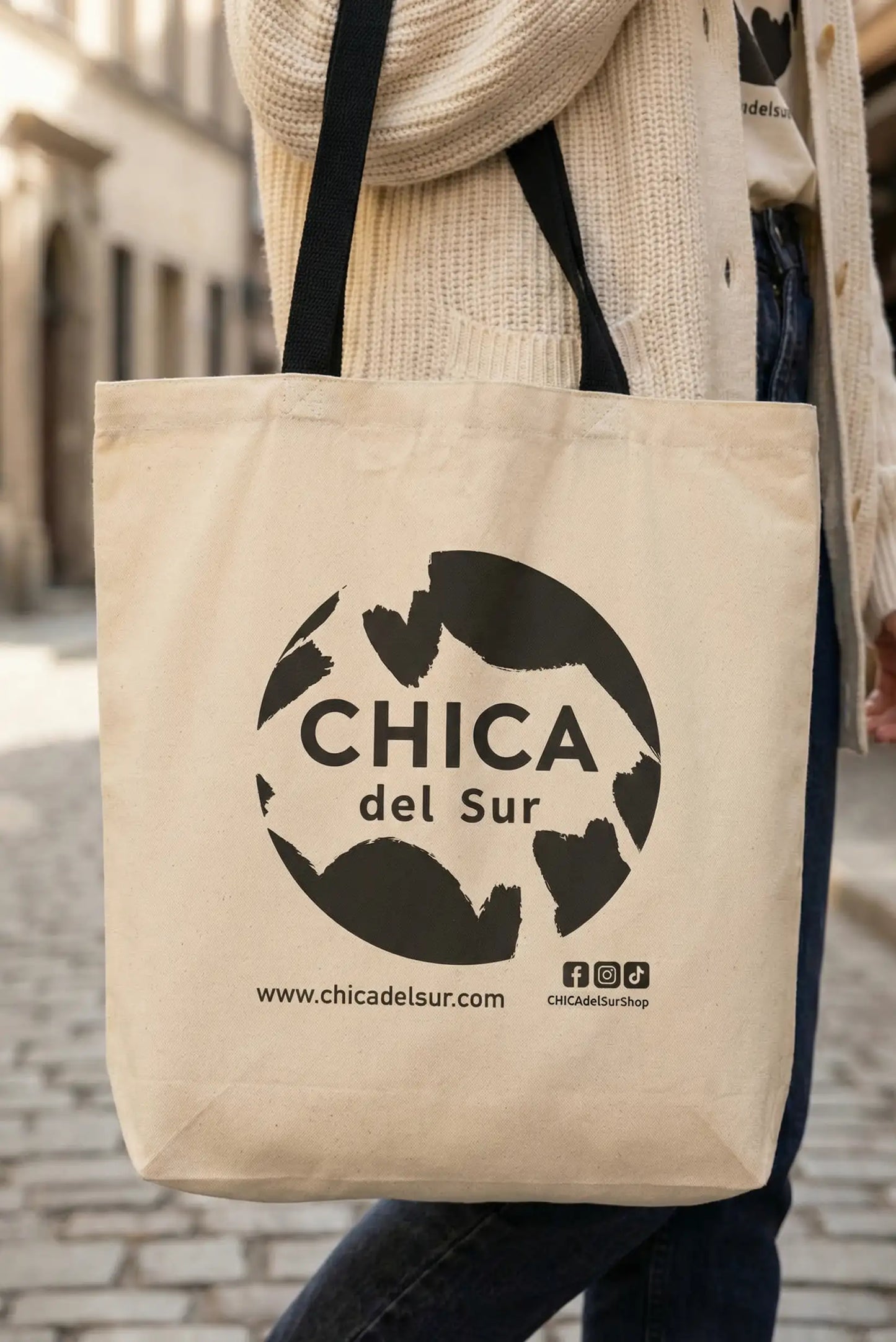 Bolsa de tela beige con el logo CHICA del Sur, moda femenina casual, en calle empedrada.