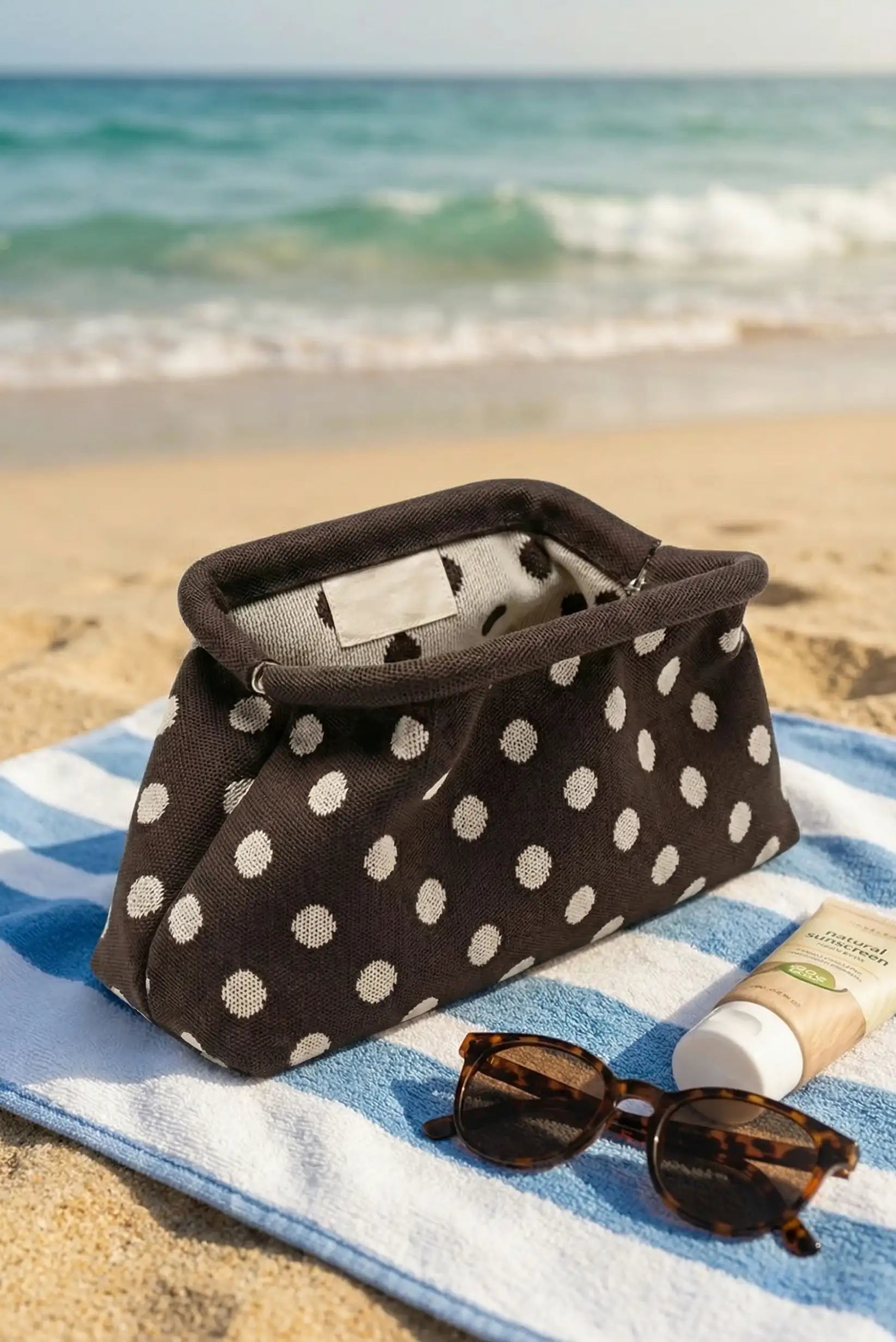 Bolso clutch marrón con topos beige sobre toalla en la playa, junto a gafas y crema solar.