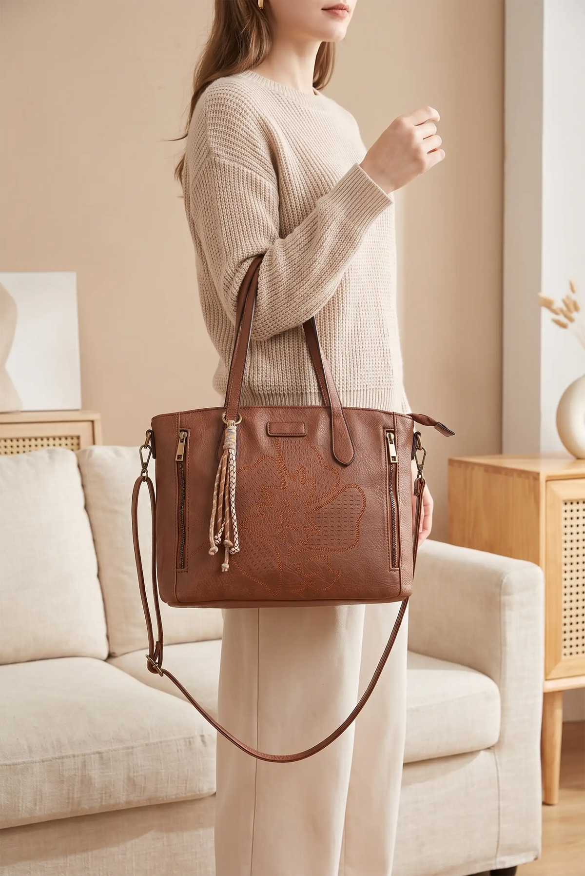 Mujer con jersey beige sostiene un bolso de mano marrón grabado con correa ajustable.