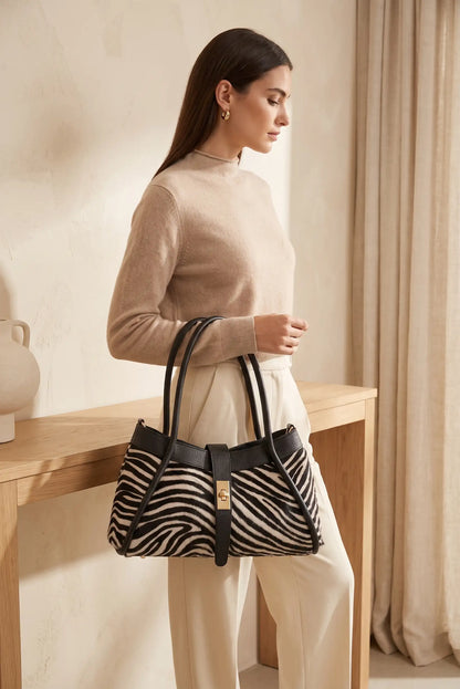 Mujer con jersey beige y pantalón blanco sosteniendo un bolso de CHICA del Sur con estampado cebra.