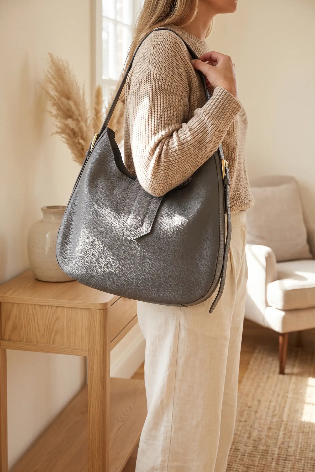 Mujer con bolso hobo de piel gris, jersey beige y pantalones blancos en interior luminoso.