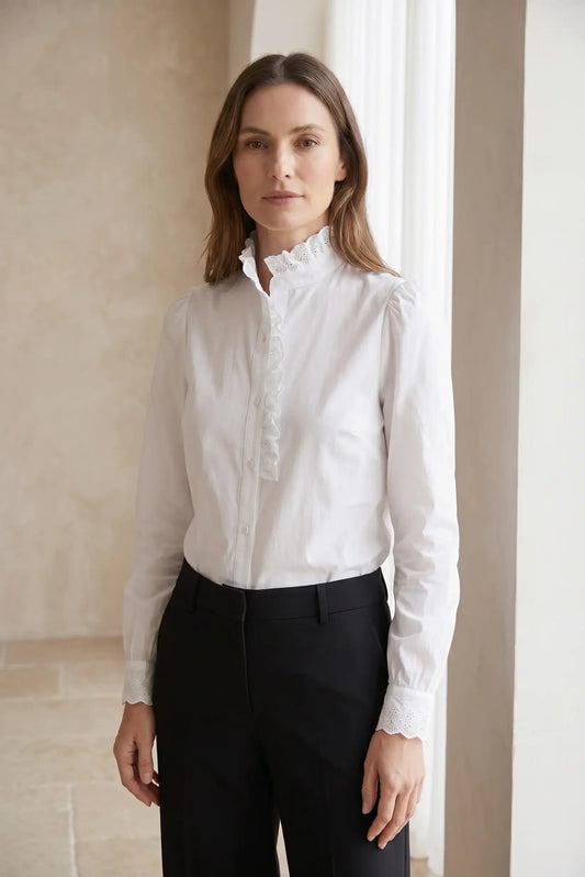 Mujer con camisa blanca de manga larga, cuello alto con volantes y pantalón negro elegante.