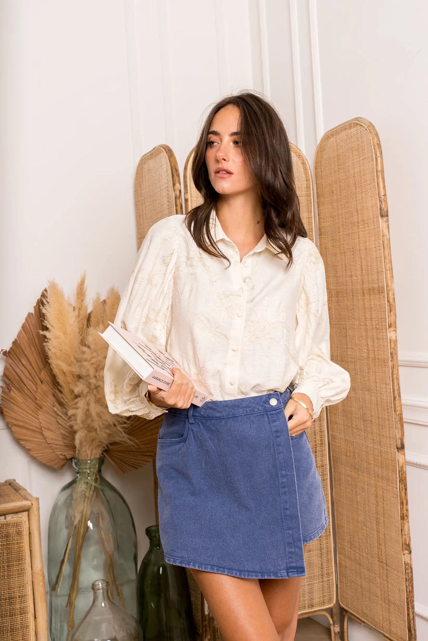 Mujer con blusa beige de manga larga abullonada y falda azul, sosteniendo libro, fondo con plantas secas