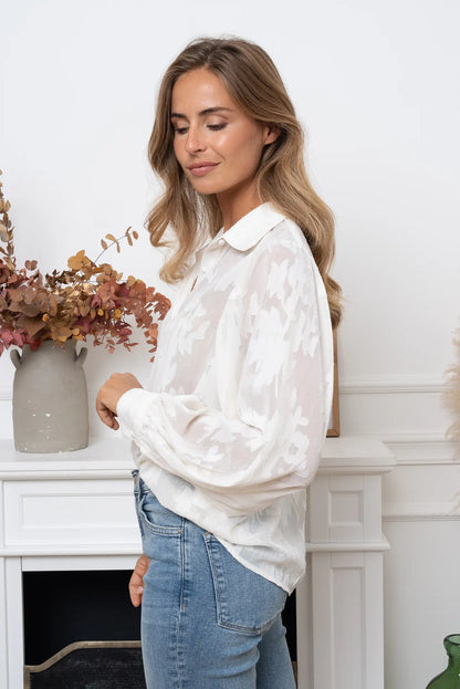 Mujer con blusa blanca roto estampada y manga larga posando de perfil en interior minimalista