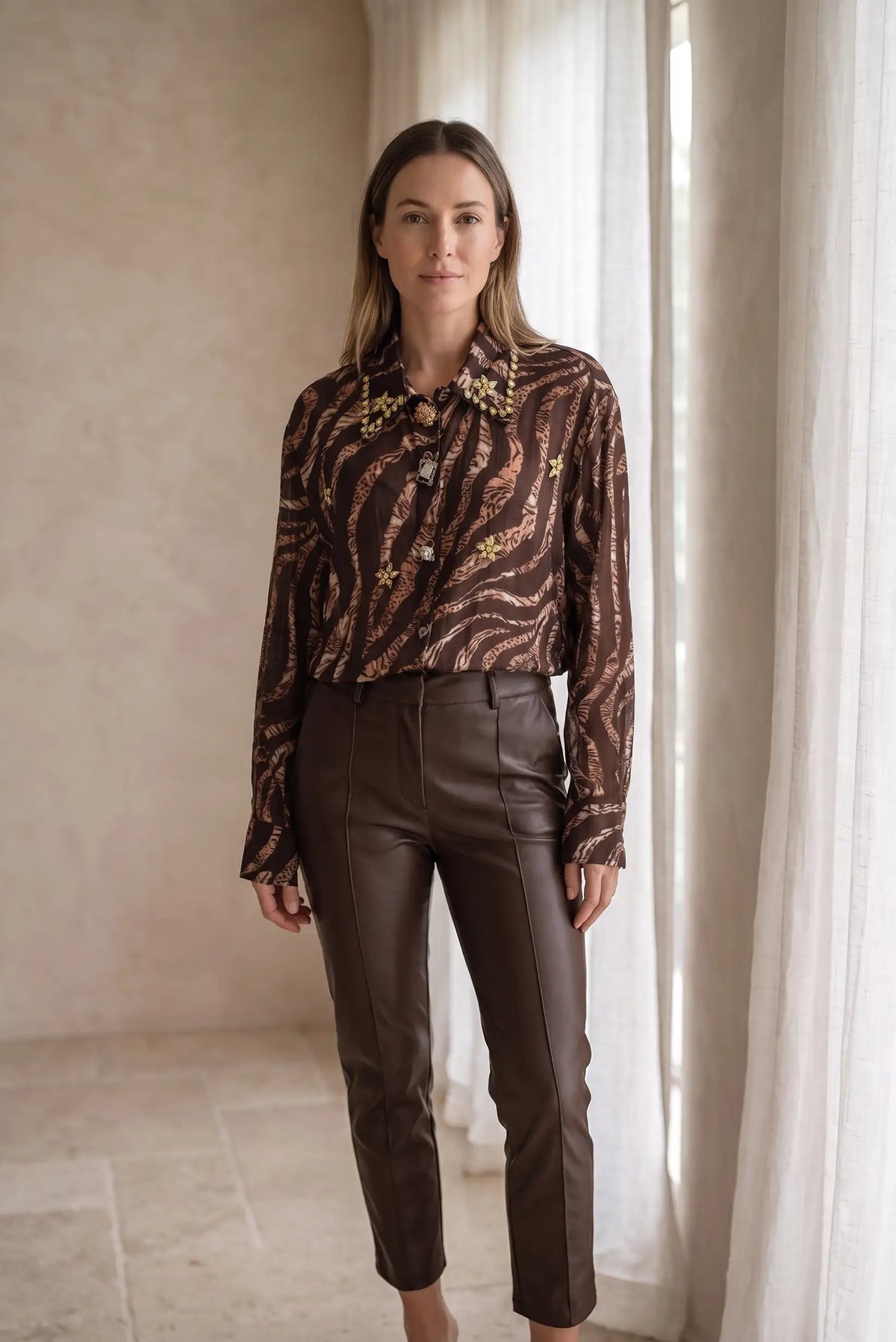 Mujer vistiendo camisa animal print marrón con pedrería dorada y pantalones de cuero marrón.