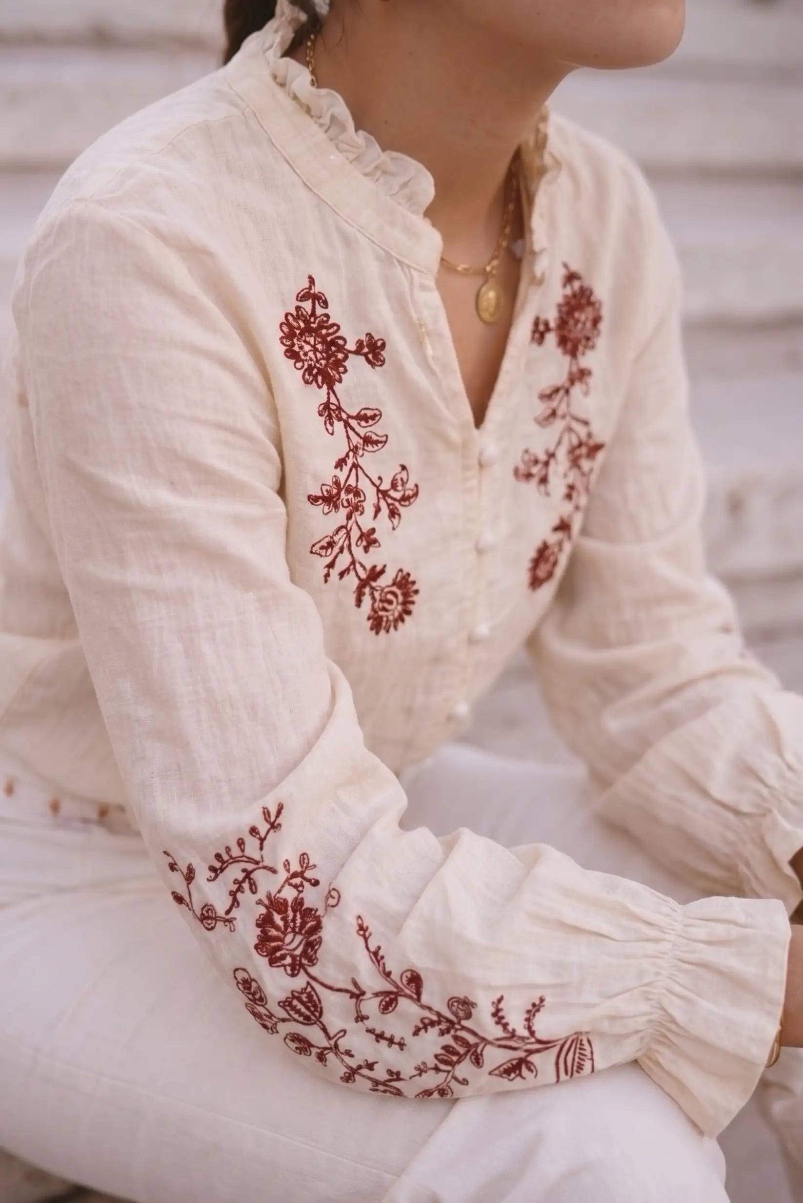 Blusa blanca con bordado floral rojo, cuello fruncido y mangas largas, ideal para looks bohemios y elegantes.
