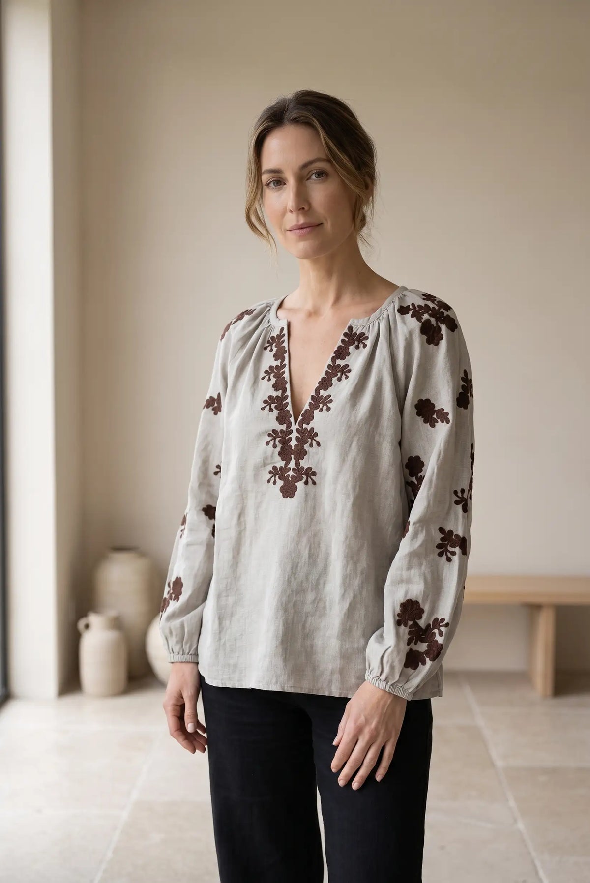 Mujer vistiendo blusa de lino beige con bordados florales marrones y pantalón negro.