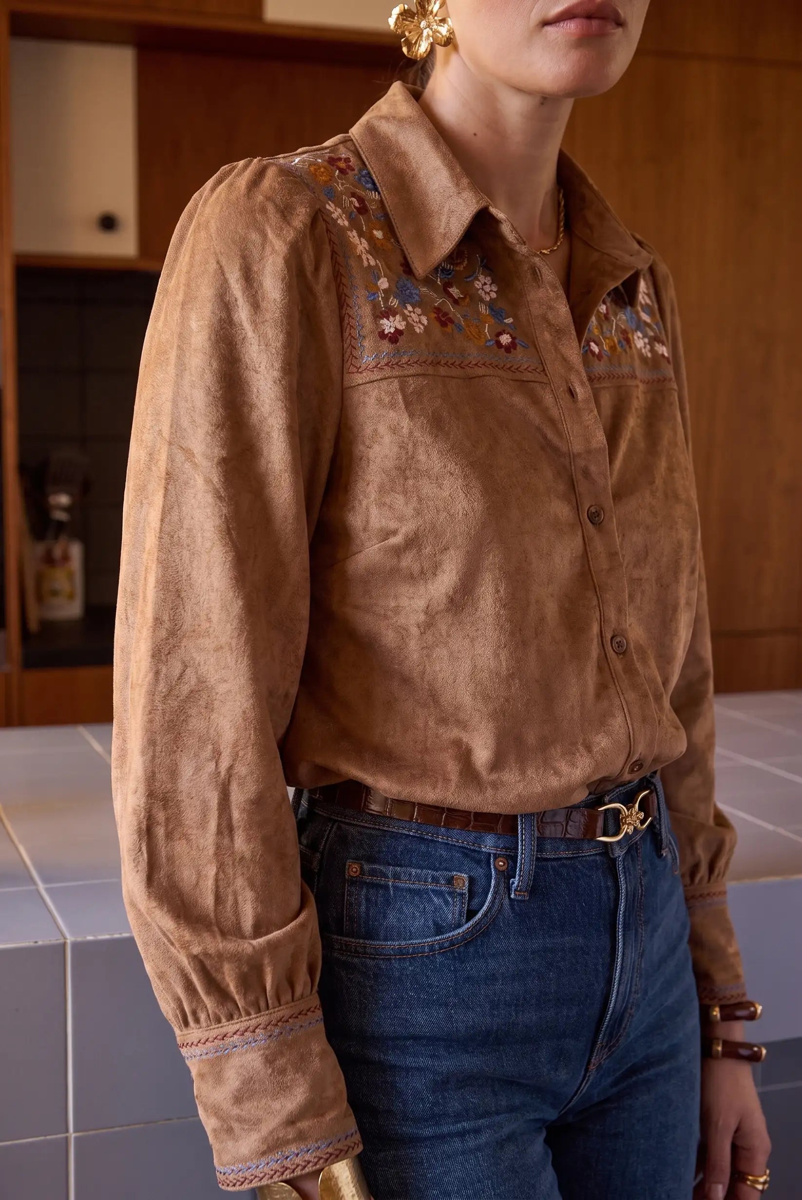 Camisa marrón de ante con bordados florales, manga larga y cuello, combinada con jeans azules y cinturón marrón.