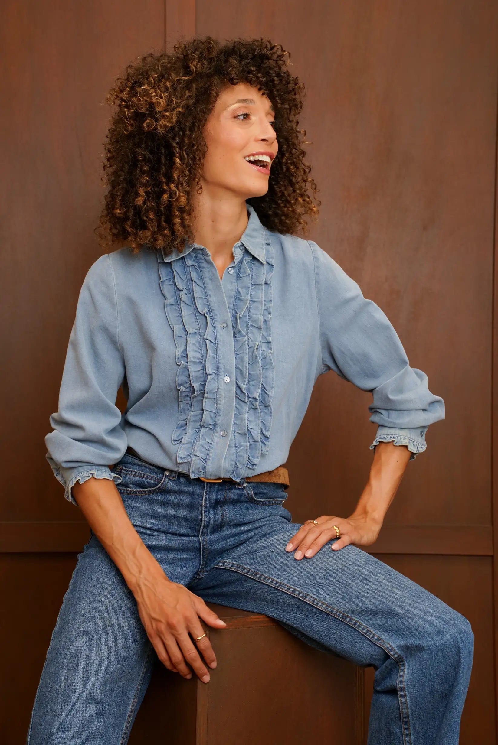 Mujer con camisa denim azul con volantes y vaqueros azules sobre fondo marrón.