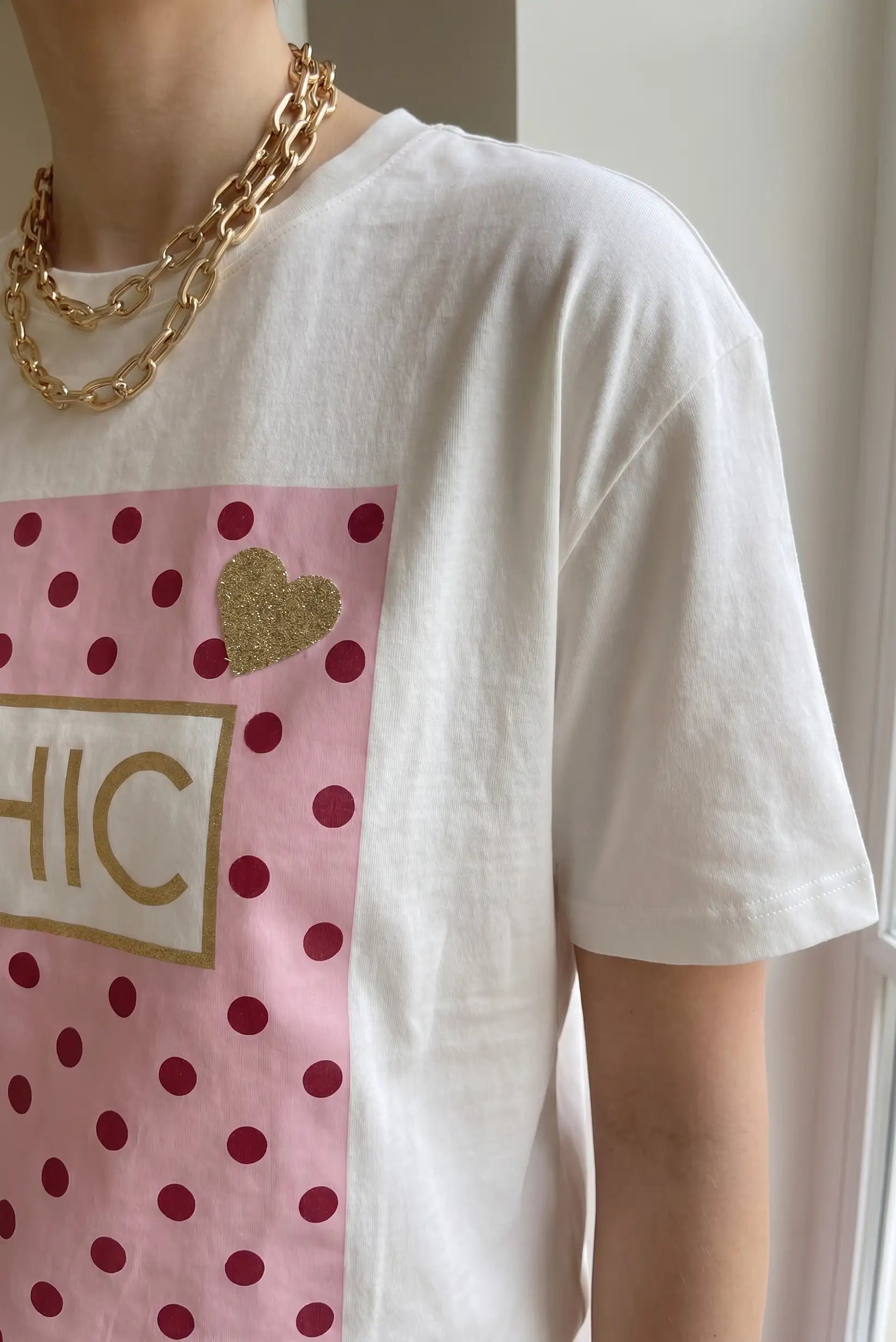 Camiseta blanca de mujer con estampado rosa de lunares y corazón dorado, estilo casual chic.