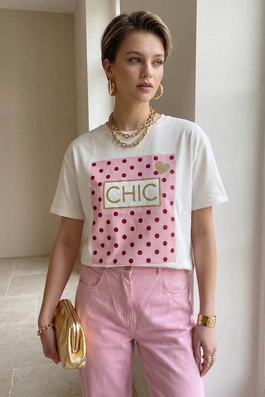 Mujer con camiseta blanca de manga corta con estampado rosa de topos y palabra CHIC, pantalón rosa y accesorios dorados