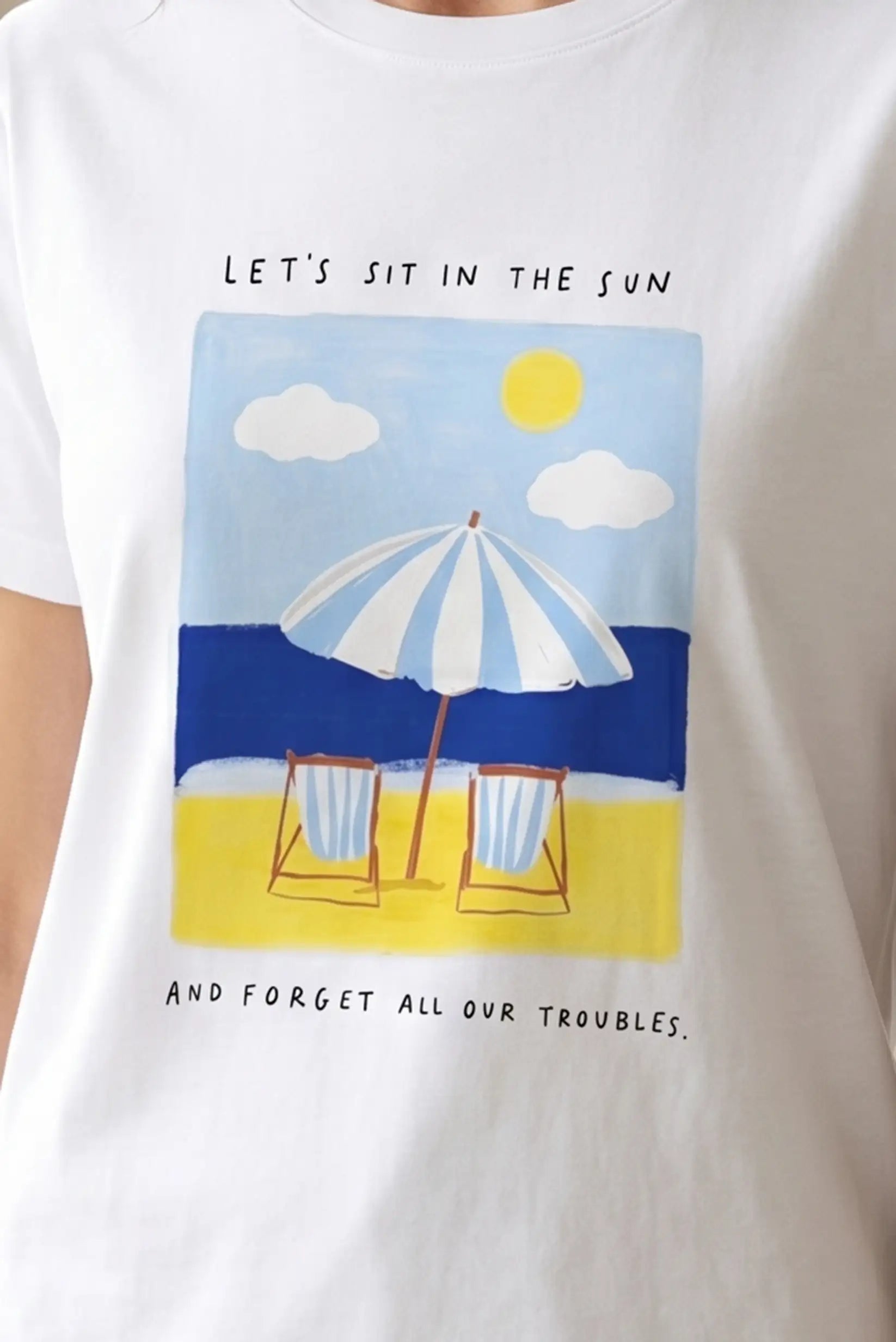 Camiseta blanca mujer con estampado playa, sombrilla azul, sillas y sol veraniego.