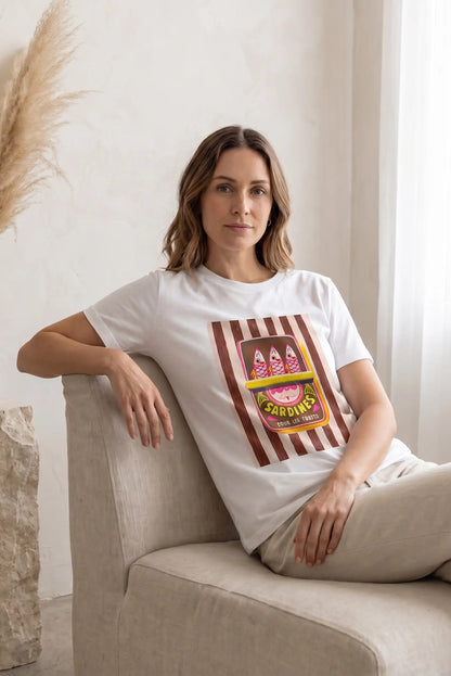 Mujer con camiseta blanca de manga corta estampada de sardinas coloridas, sentada en sofá beige.