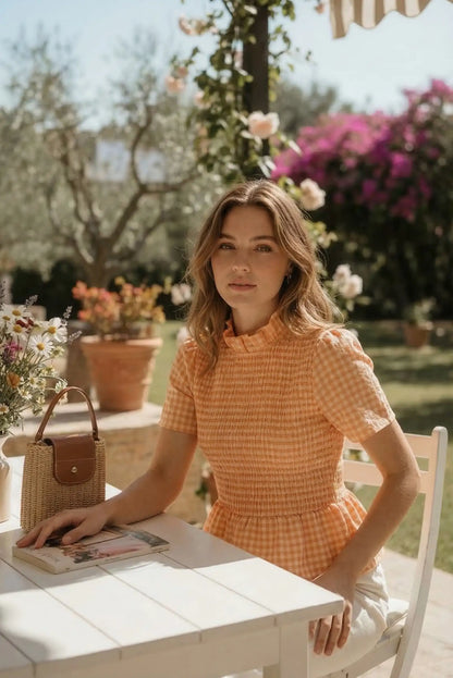 Mujer con blusa fruncida de cuadros naranja, sentada en jardín, bolso de mimbre sobre mesa.