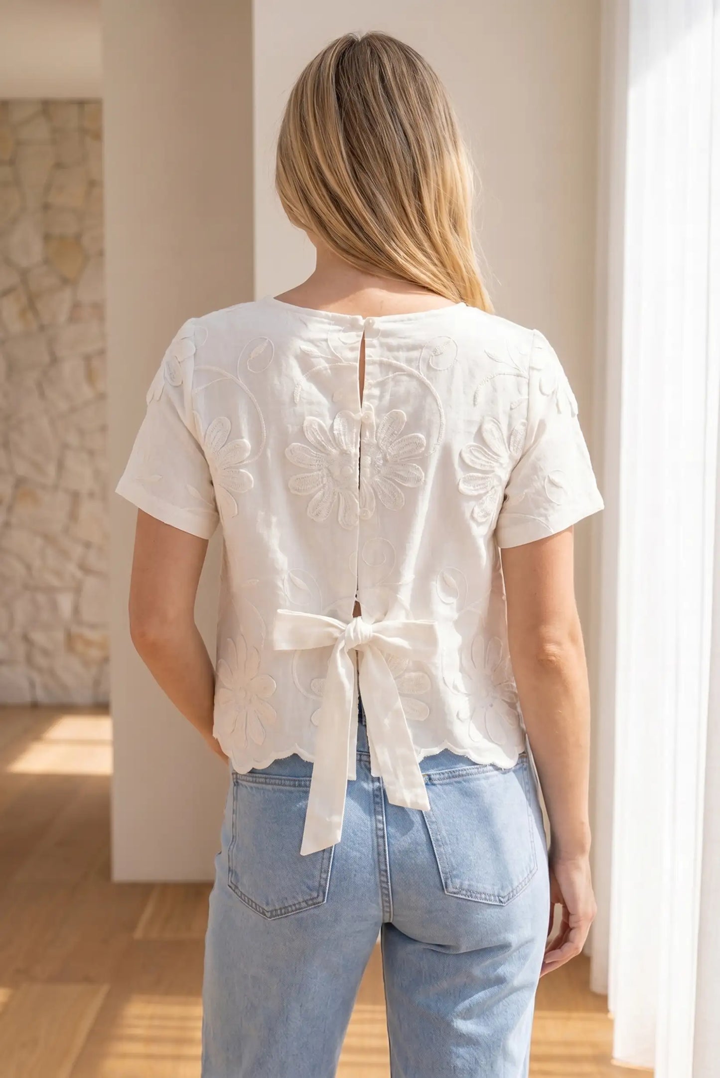 Mujer con camiseta blanca de manga corta, flores bordadas y lazo en la espalda, estilo casual.