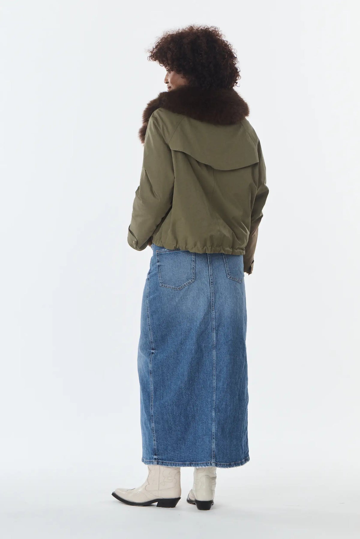 Mujer de espaldas con chaqueta verde oliva con cuello de pelo, falda vaquera larga y botas beige.