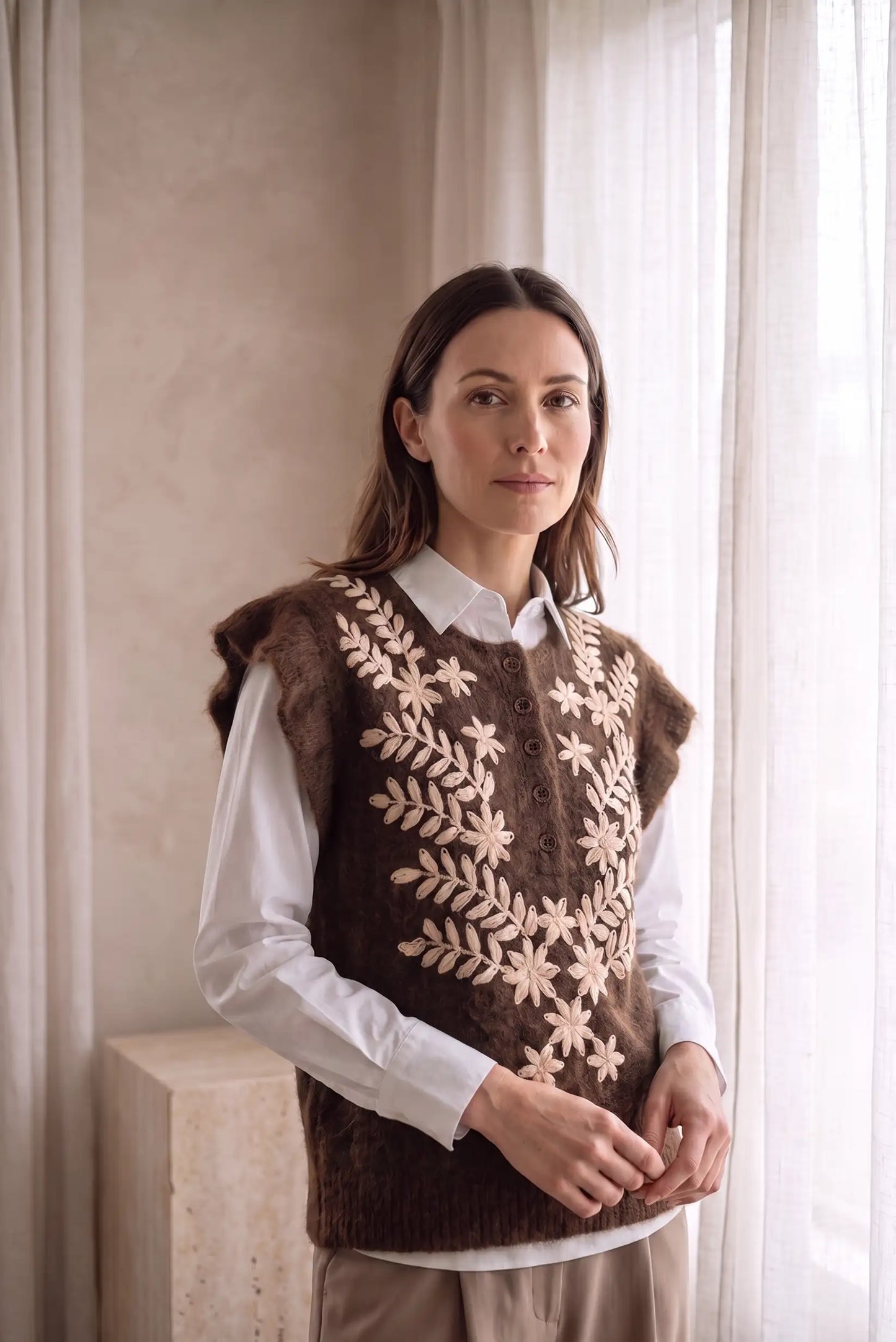 Mujer con chaleco marrón bordado con flores beige sobre camisa blanca.