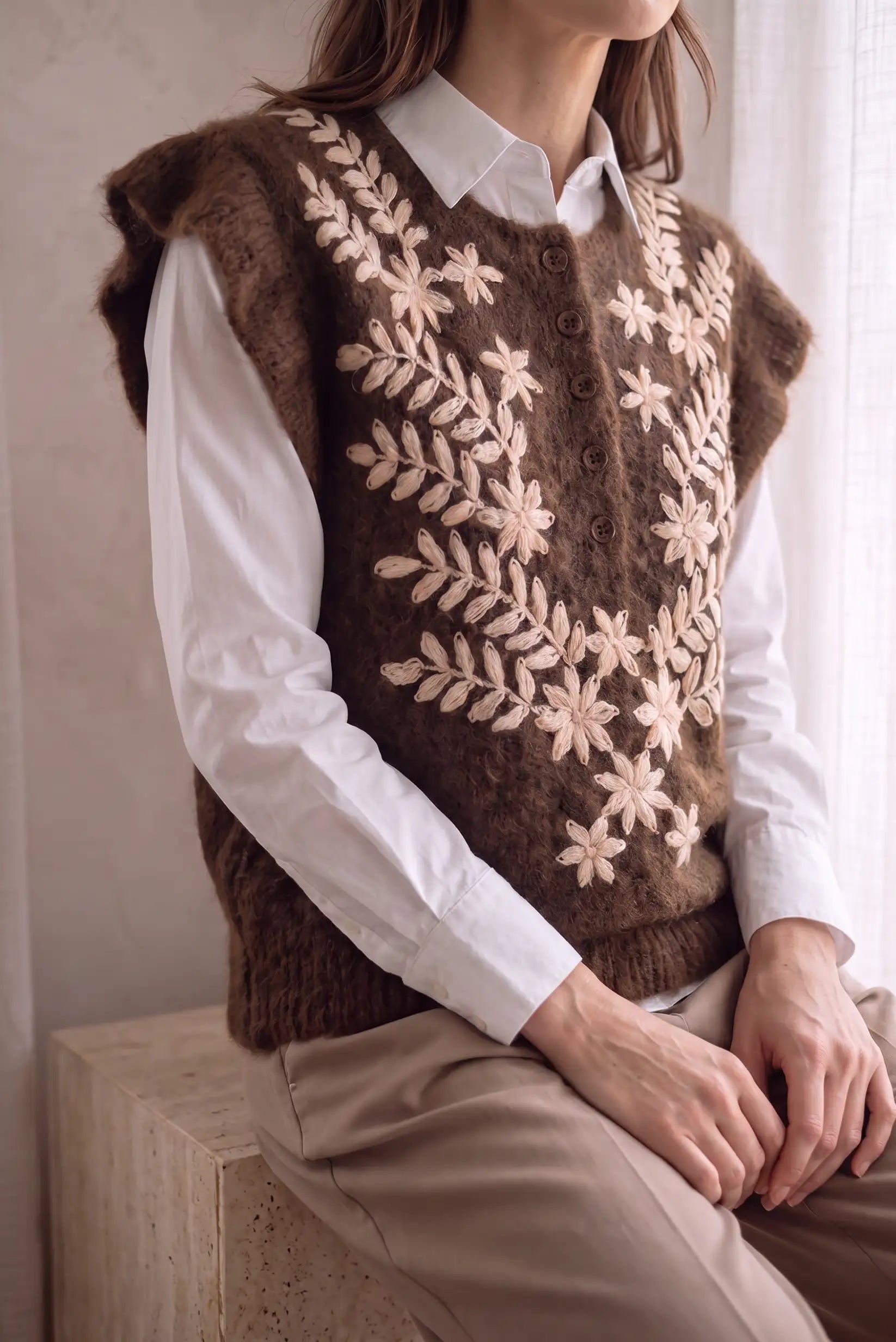 Chaleco de punto marrón con bordados florales beige sobre camisa blanca y pantalón beige.